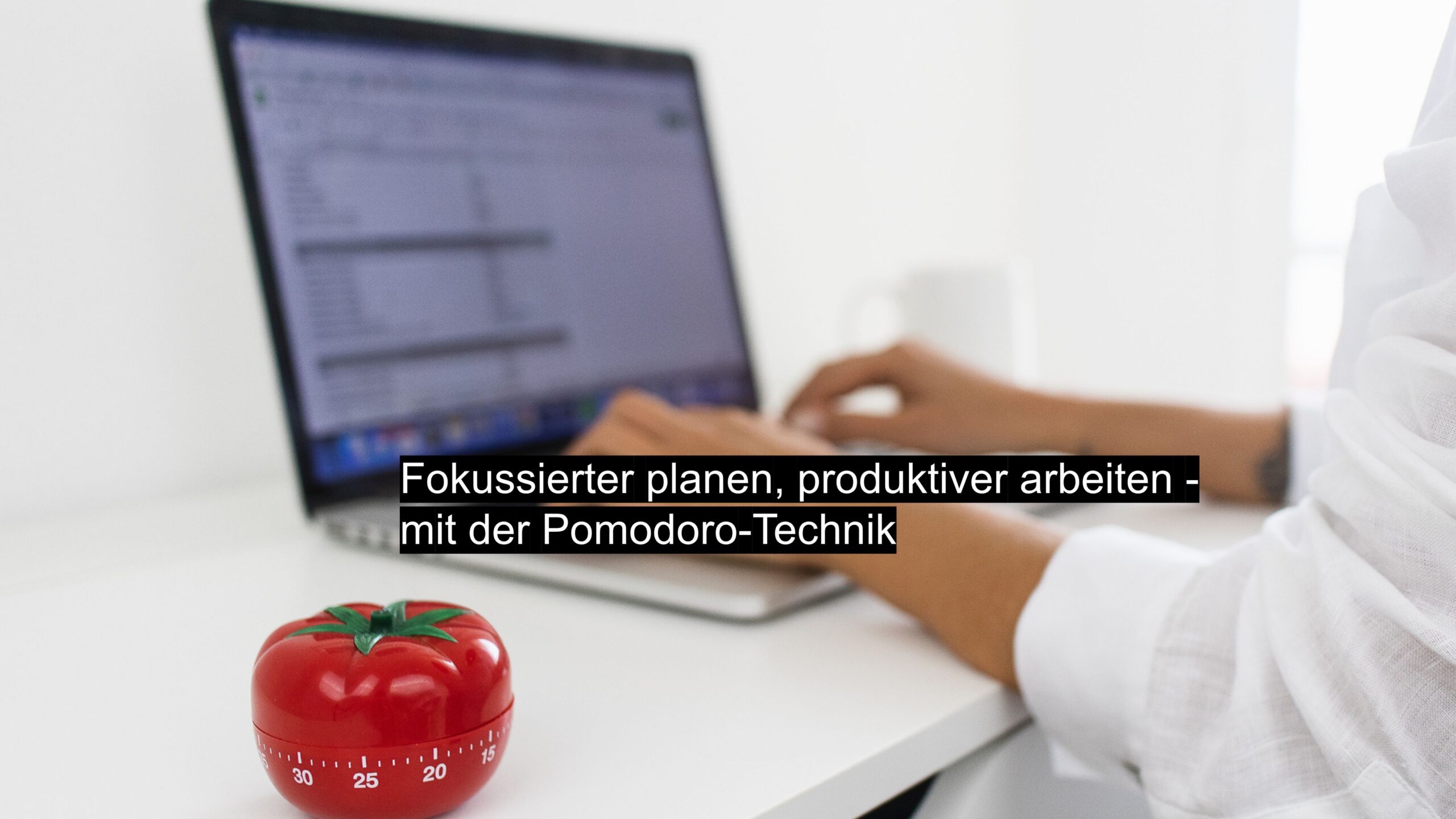 Pomodoro-Timer in Tomatenform auf Schreibtisch, im Hintergrund Person beim Arbeiten am Laptop – Fokus und Zeitmanagement mit der Pomodoro-Technik.